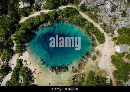 Aus der Vogelperspektive auf ein faszinierendes, tiefblaues Sinkloch, das sich im Kontrast zur umgebenden trockenen Landschaft und üppigen grünen Vegetation in Cetina, Kroatien, befindet. Stockfoto