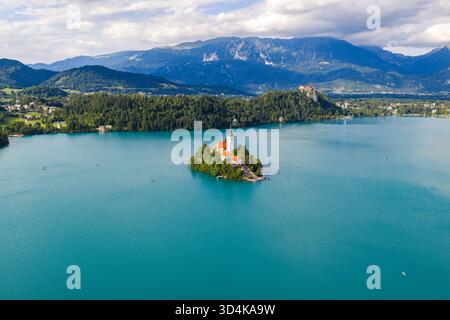 Blick aus der Vogelperspektive auf die berühmte Kirche auf Bled Island, die vor dem türkisfarbenen See schimmert, eingerahmt von grünen Wäldern und dem fernen Bled Castle, Bled, Ra Stockfoto