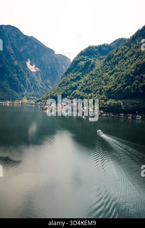 Aus der Vogelperspektive eines Bootes, das durch das ruhige, reflektierende Wasser eines Sees gleitet, umgeben von grünen Bergen und dem charmanten Dorf Hallstatt, Upper Stockfoto