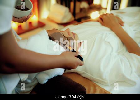 In der ruhigen Atmosphäre des Spa-Salons verwöhnt die Kundin ihre Gäste mit einer Gesichtscreme mit Holzkohle und einer warmen Kerze. Gesichtshaut Stockfoto