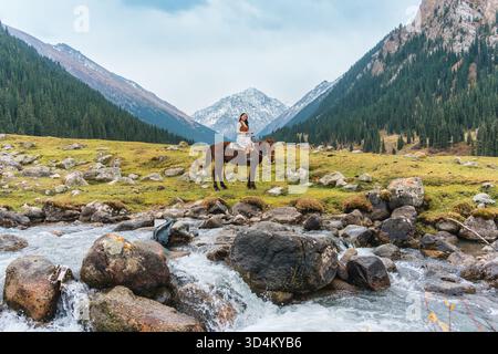 Schöne asiatische Frau reitet Pferd im malerischen Bergtal von Altyn Arashan mit Fluss und schneebedeckten Berg in Kirgisistan Stockfoto