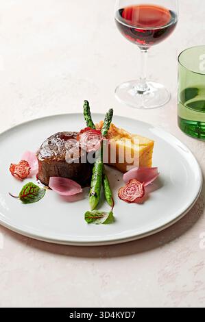 Augenfilet mit Kartoffelgratin, verkohltem Spargel, shiraz jus, eingelegten Schalotten und Rote-Bete-Chips auf rosa Oberfläche mit Wein- und Wassergläsern Stockfoto
