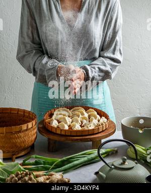 Weibliche Schafe, die ein Bambustablett mit ungekochten Gyoza-Knödeln hält, Bambusdampfer. Teekanne teal, frische grüne Zwiebeln und Pilze, asiatische Küche Stockfoto