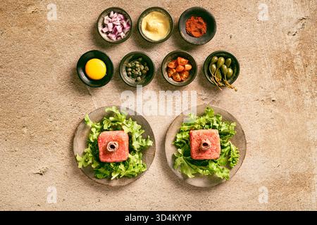 Flaches Lagen von frisch zubereitetem rohem Rindfleischtartar in zwei separaten Tellern mit Salat und Sardellen, die verzehrt werden können, mit verschiedenen Gewürzsorten und s Stockfoto