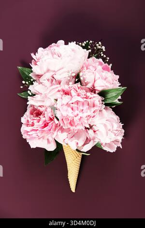 Künstlerisches Arrangement aus rosa Pfingstrosen und Grün in einem Waffelkegel vor einem üppigen burgunderroten Hintergrund. Stockfoto