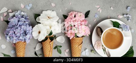 Kreative flache Ladefläche mit blumengefüllten Waffelkegeln und einer Tasse Kaffee, umgeben von verstreuten Blütenblättern auf einer grauen strukturierten Oberfläche. Stockfoto