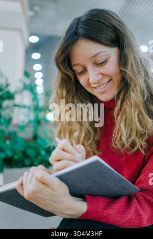 Junge Frau lächelt, während sie sich darauf konzentriert, in einem persönlichen Notizbuch zu schreiben, Gedanken zu erfassen, Kreativität zu verfolgen und ihr Wissen während eines konzentrierten Studien- oder Journalsitzens zu erweitern Stockfoto