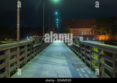 Eine lange Holzbrücke mit Straßenlaternen führt nachts zur Burg Malbork in Nordpolen. Warme Wände und Dächer reflektieren den Fluss Nogat, eingerahmt von Stockfoto