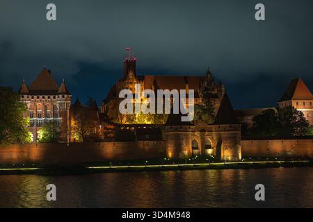 Warme Scheinwerfer beleuchten die Burg Malbork am Fluss Nogat in Polen bei Nacht. Zinnenverkleidete Mauern, spitze Türme und das bogenförmige Torhaus reflektieren das Wasser Stockfoto