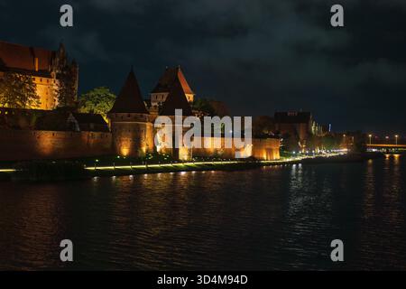 Rote Backsteinmauern und konische Türme leuchten nach der Dämmerung mit warmen Lichtern. Auf dem Fluss Nogat tummeln sich Reflexionen, während die riesige gotische Festung die s dominiert Stockfoto