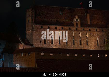 Schloss Malbork in Polen bei Nacht, gotische Mauern und Türme beleuchtet von warmen Lichtern, Lanzettenfenster und Mauerwerk sichtbar, rote Flagge auf dem Kamm, Textur und sc Stockfoto
