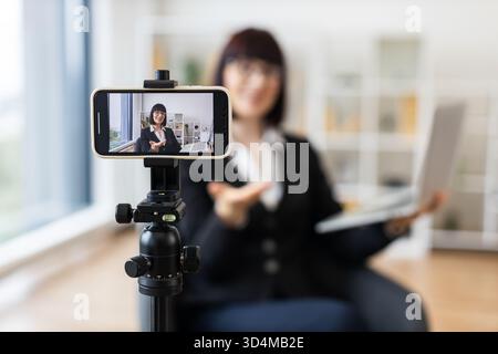 Kaukasische Frau in Geschäftskleidung, die sich auf dem Smartphone aufzeichnet und geschäftliche Themen im Büroumfeld diskutiert. Konzept des Video-Bloggens, online Stockfoto