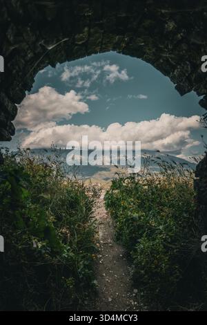 Blick durch den alten Steinbogen in die Berglandschaft mit wilden Pflanzen, blauem Himmel und dramatischen Wolken. Stockfoto