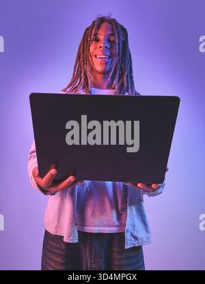 Junger Mann mit lächelnden Dreadlocks, der einen offenen Laptop hält, beleuchtet von neonviolettem Licht Stockfoto