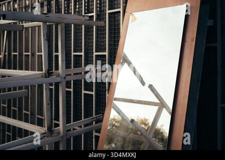 Großer alter Spiegel, der sich im Freien gegen eine Gitterstruktur aus Metall lehnt und Himmel und geometrische Metallstäbe in einer industriellen städtischen Umgebung reflektiert. Selektiv Stockfoto