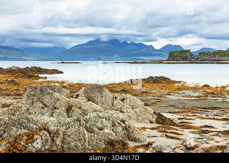 Felsige Küste mit Algen- und Gezeitenbecken mit Blick auf die Ruinen von Dunscaith Castle und die Cuillin Mountains auf der Isle of Skye, Schottland, reflektierend über den Fluss Stockfoto