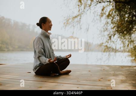 Eine Koreanerin sitzt friedlich auf einer hölzernen Plattform in der Nähe des Sees und übt Meditation, während die Sonne untergeht, umgeben von der Natur. Stockfoto