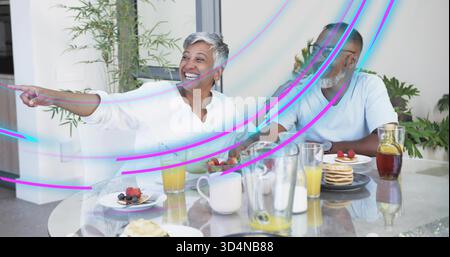 Lachende Frau in weisser Bluse, Mann in hellem T-Shirt, der zuhause zusieht, Pfannkuchen auf dem Tisch Stockfoto