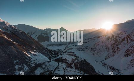 Atemberaubende Aussicht auf eine gewundene Straße, die sich durch ein schneebedecktes Tal schlängelt, eingebettet zwischen hoch aufragenden, sonnengeküssten Gipfeln, während die Sonne unter dem Horizont untergeht und ein warmes, goldenes Leuchten erzeugt Stockfoto