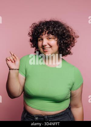 Eine junge Person mit lockigen Haaren trägt ein grünes, verkürztes Hemd und drückt Freude aus, indem sie lächelt und mit dem Finger zeigt. Der leuchtend pinkfarbene Hintergrund fügt hinzu Stockfoto