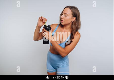 Eine Frau mit blauem sportlichem Outfit konzentriert sich darauf, eine schwarze Shaker-Flasche zu ergänzen. Sie steht in einem normalen Studio und zeigt ihre Fitnes Stockfoto