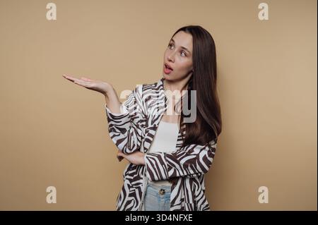 Eine Frau mit langen Haaren steht mit erhobener Hand und drückt Überraschung oder Neugier aus. Sie trägt eine gemusterte Jacke und eine lässige Jeans, die wieder Set ist Stockfoto