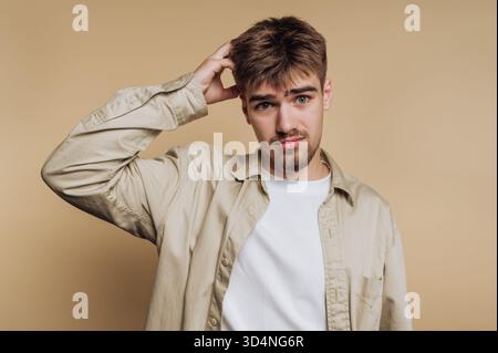 Ein junger Mann steht vor einem schlichten beigen Hintergrund, sieht verwirrt aus und kratzt sich den Kopf. Sein lässiges Outfit vermittelt eine entspannte, ansprechende Atmosphäre. Stockfoto