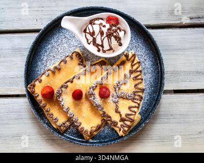 Blick von oben auf Pfannkuchen mit Schokoladenmuster, Erdbeeren und Eis in separater Schüssel auf Keramikplatte auf Holztisch Stockfoto