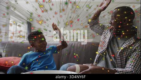 High-Fiving Dad und Sohn auf grauem Sofa zu Hause, kariertes Hemd, blaues T-Stück, Football-and-Bowl, Konfetti Stockfoto