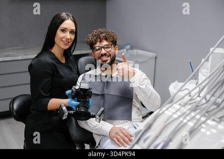 Zahnarzt hält eine Kamera mit Ringlicht, fotografiert einen lächelnden männlichen Patienten, der in einer modernen Klinik den Daumen nach oben zeigt Stockfoto