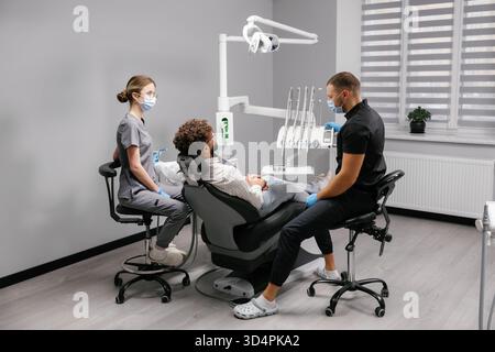 Mann mit Brille und lockigem Haar lächelt in einem Zahnarztstuhl, während Zahnarzt seine Zähne überprüft Stockfoto