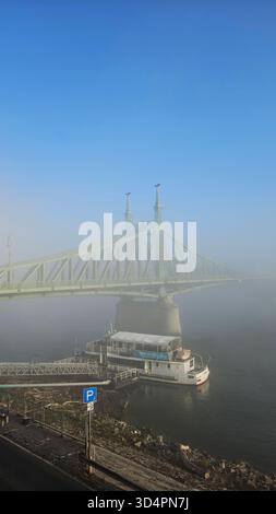 Liberty Bridge und Bootstour auf dem Dock an einem nebeligen, aber sonnigen Herbstmorgen über der Donau in Budapest, Ungarn Stockfoto