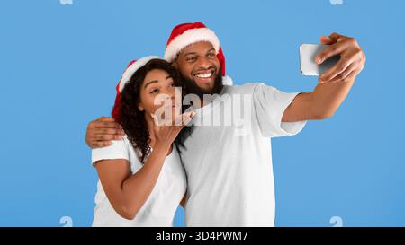 Verspielte afroamerikanische Ehepartner in Santa-Hüten machen Selfie auf dem Smartphone, Frau bläst Luftkuss auf die Handykamera Stockfoto