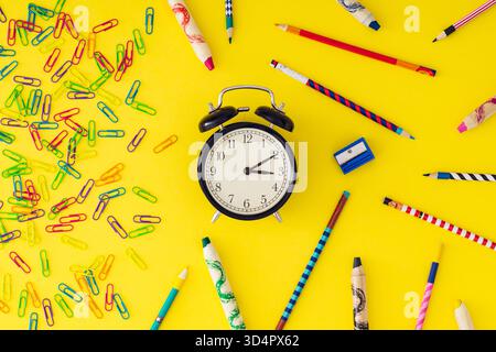 Bunte Komposition für den Schulbeginn mit Retro-Wecker, Büroklammern, Wachsmalstiften und Buntstiften auf gelbem Hintergrund. Stockfoto