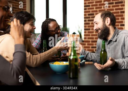 Bärtiger weißer Mann, der nach einem Jenga-Spiel mit multiethnischen Freunden urkomische Kindheitsgeschichte erzählt. Gruppe fröhlicher Begleiter, die sich bei Getränken, Snacks und lustigen Witzen im gemauerten Raum verbündet. Stockfoto