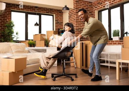 Fröhliches junges Paar, das den Umzugstag feiert, als Frau, die Box hält, von einem afroamerikanischen Ehemann in einer neuen Wohnung auf einen Stuhl geschoben wird. Ich war begeistert von verschiedenen verheirateten Personen, die Spaß in einem unmöblierten Zimmer hatten. Stockfoto