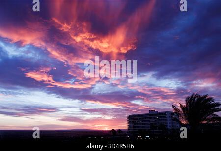 Dramatischer, lebendiger Sonnenuntergangshimmel mit violetten und orangen Wolken über einem modernen Strandhotel und Silhouettenpalmen Can pastilla mallorca spanien Stockfoto