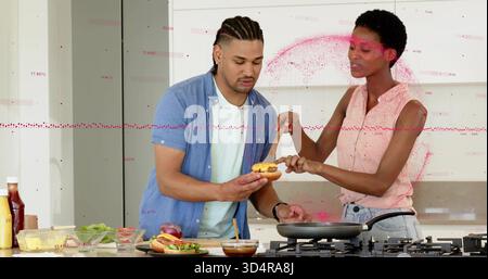 Ein Paar, das legere Hemden und Jeans trägt und Brötchen und Löffelsauce in der Küche hält Stockfoto