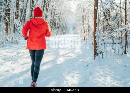 Trailläuferin in leuchtend roter, winddichter Jacke, die an einem sonnigen, frostigen Tag einen malerischen, verschneiten Waldweg joggen lässt. Sportlich aktive Menschen und Winter Stockfoto