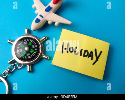 Dieses Bild zeigt ein Reisethema mit einem Spielzeugflugzeug, einem Kompass-Schlüsselanhänger und einer Haftnotiz mit dem Wort Holiday. Stockfoto