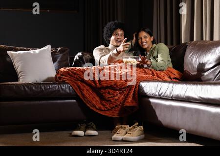 Diverse Paare kuscheln sich zu Hause auf der Couch, teilen Popcorn und rostfarbene Überwurfdecke Stockfoto