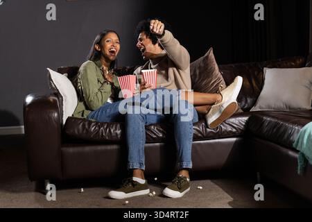 Lachende, vielseitige Paare, die im Wohnzimmer auf einer Ledercouch sitzen und Popcorn-Wannen halten Stockfoto