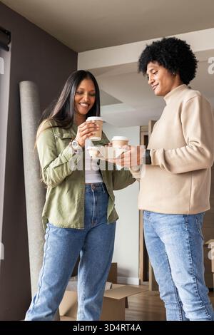 Mit Kaffeetassen zum Mitnehmen, diverses Paar im Haus, mit Umzugskartons, gerolltem Teppich Stockfoto