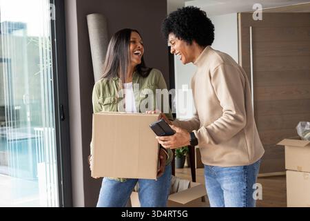 Lachende Paare, die Umzugskartons in einem modernen Eingangsbereich auspacken, mit Smartphone in der Hand Stockfoto