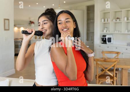 Verschiedene Freundinnen singen Karaoke in der modernen Küche, Essbereich mit Mikrofonen Stockfoto Verschiedene Freundinnen singen Karaoke in der modernen Küche, Essbereich mit Mikrofonen Stockfoto
