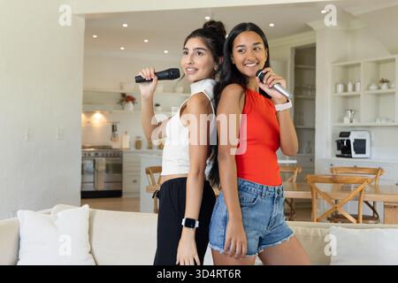 Sie singen verschiedene weibliche Freunde, die zu Hause auf dem Sofa stehen, mit drahtlosen Mikrofonen Stockfoto Sie singen verschiedene weibliche Freunde, die zu Hause auf dem Sofa stehen, mit drahtlosen Mikrofonen Stockfoto