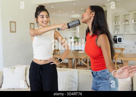 Verschiedene Freundinnen singen, die Karaoke im offenen Wohnzimmer mit Haarbürste spielen Stockfoto Verschiedene Freundinnen singen, die Karaoke im offenen Wohnzimmer mit Haarbürste spielen Stockfoto