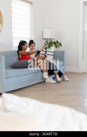 Verschiedene Freundinnen sitzen, lächeln und zeigen auf dem hellblauen Sofa mit Kissen auf das Smartphone Stockfoto