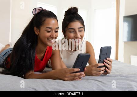 Lächelnde, vielfältige Freundinnen, die Seite an Seite auf dem Bett liegen, im hellen Schlafzimmer, mit Smartphones Stockfoto