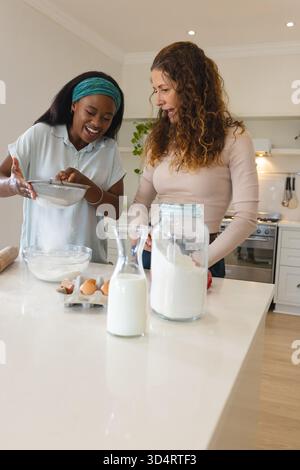 Verschiedene Freundinnen sieben Mehl und Lächeln in der modernen Küche, mit einer Rührschüssel Stockfoto